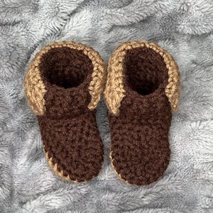 May include: A pair of handmade crochet baby booties. The booties are brown with a tan trim around the top. The booties are made of soft yarn and are likely designed for comfort and warmth. The background is a soft, gray, textured fabric.