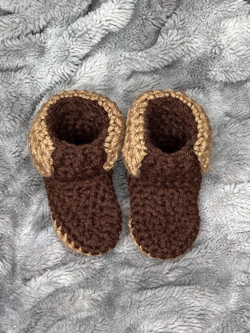 May include: A pair of handmade crochet baby booties. The booties are brown with a tan trim around the top. The booties are made of soft yarn and are likely designed for comfort and warmth. The background is a soft, gray, textured fabric.