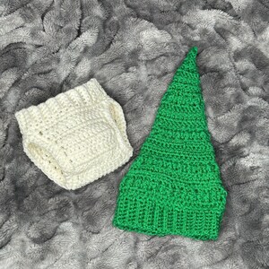May include: A handmade crochet set featuring a green pointed hat and a pair of off-white diaper covers. The items are made of soft yarn and are displayed on a gray, textured surface. The set is likely for a baby or infant.