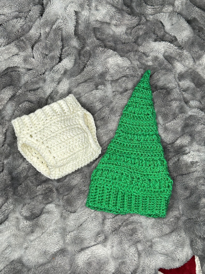 May include: A handmade crochet set featuring a green pointed hat and a pair of off-white diaper covers. The items are made of soft yarn and are displayed on a gray, textured surface. The set is likely for a baby or infant.