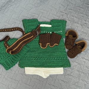 May include: A handmade crochet baby outfit in green, brown, and white. The set includes a tunic, shorts, hat, belt, wrist cuffs, and booties. The outfit is designed with a textured stitch and is laid out on a gray surface.