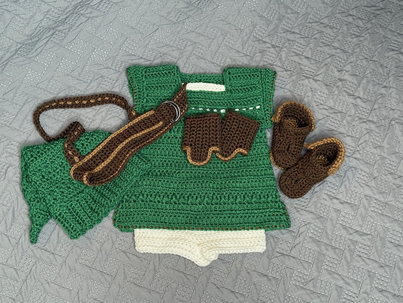 May include: A handmade crochet baby outfit in green, brown, and white. The set includes a tunic, shorts, hat, belt, wrist cuffs, and booties. The outfit is designed with a textured stitch and is laid out on a gray surface.