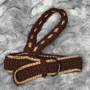 May include: A handmade crochet belt in shades of brown and tan. The belt features a woven design with a metal ring buckle. The belt is a unique accessory for adding a touch of handcrafted style to any outfit.