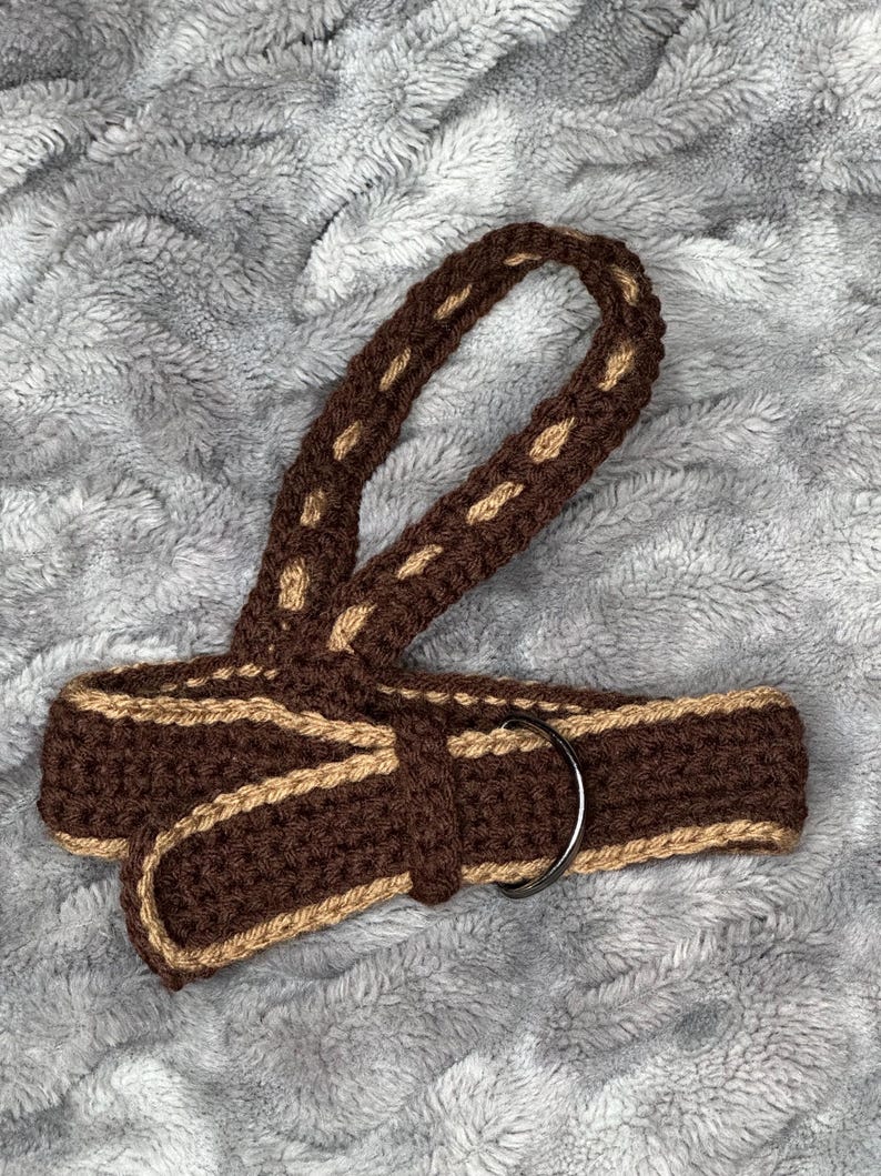 May include: A handmade crochet belt in shades of brown and tan. The belt features a woven design with a metal ring buckle. The belt is a unique accessory for adding a touch of handcrafted style to any outfit.