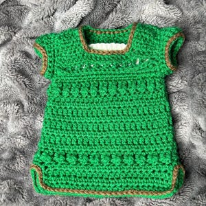 May include: A handmade green crochet top with short sleeves and a square neckline. The edges are trimmed with a brown yarn. The top is made of a textured yarn and appears to be a baby or toddler garment.