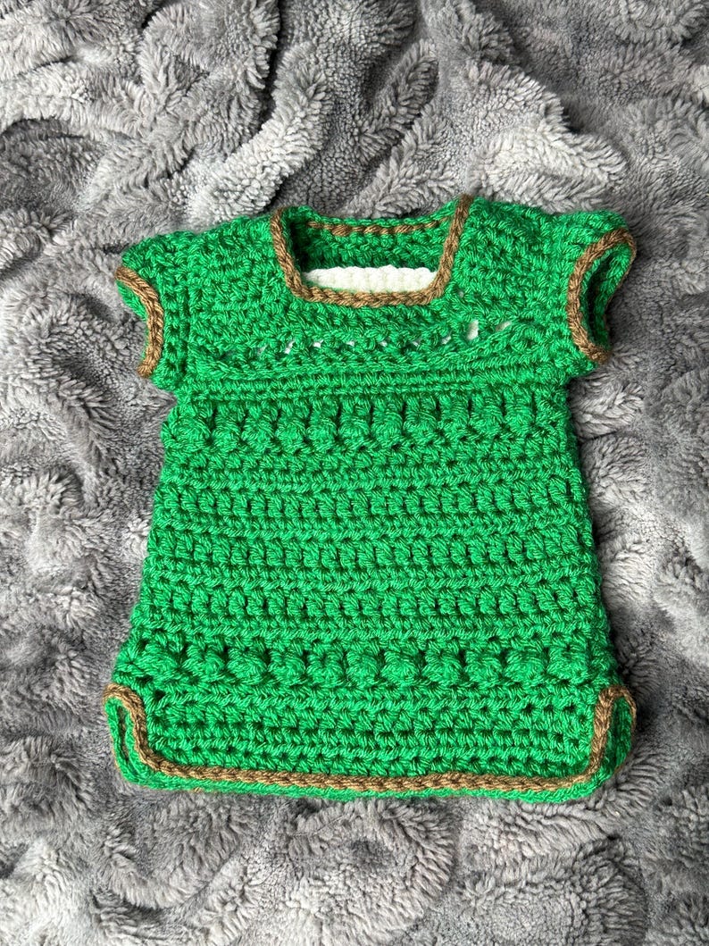 May include: A handmade green crochet top with short sleeves and a square neckline. The edges are trimmed with a brown yarn. The top is made of a textured yarn and appears to be a baby or toddler garment.