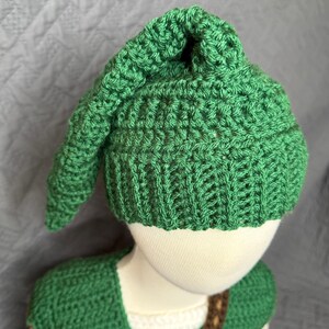 May include: A green crochet hat with a pointed top and a matching green crochet top. The hat and top are made of a textured yarn. The outfit is designed to resemble a character from a video game.
