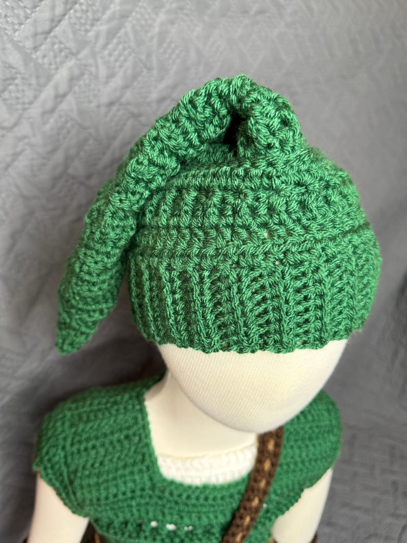 May include: A green crochet hat with a pointed top and a matching green crochet top. The hat and top are made of a textured yarn. The outfit is designed to resemble a character from a video game.
