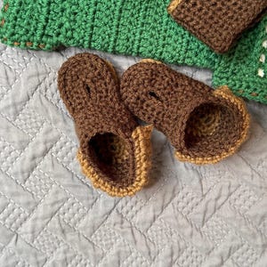 May include: A pair of handmade crochet baby booties in brown with tan trim, resting on a light gray quilted surface. A green crocheted blanket and matching brown cuff are in the background.
