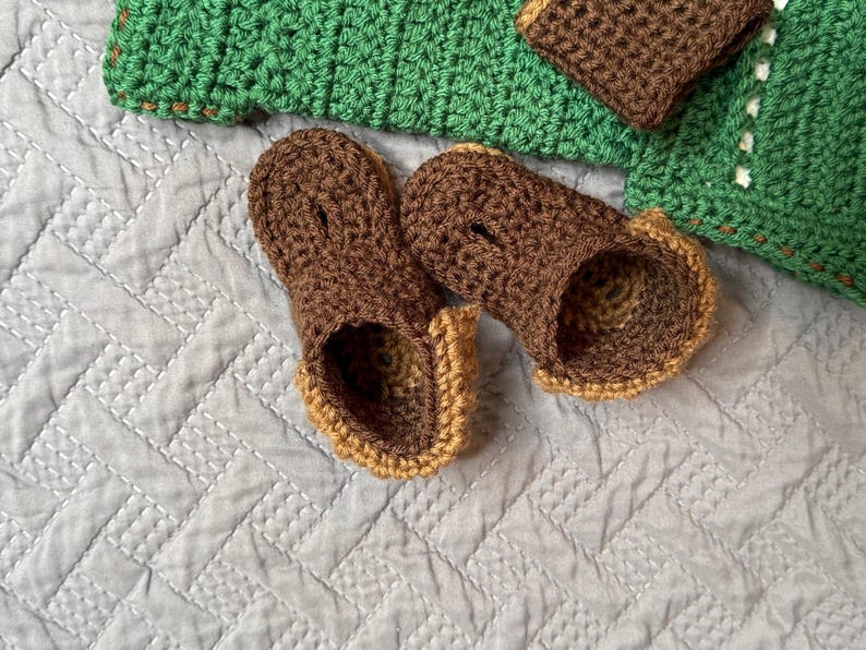 May include: A pair of handmade crochet baby booties in brown with tan trim, resting on a light gray quilted surface. A green crocheted blanket and matching brown cuff are in the background.