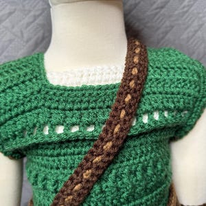 May include: A handmade green crochet top with a brown strap and accents. The top has a square neckline and a textured stitch pattern. The strap is a darker brown with lighter brown detailing. The top is designed for a child.