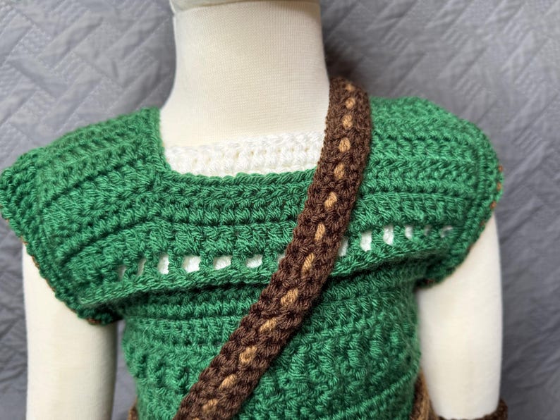 May include: A handmade green crochet top with a brown strap and accents. The top has a square neckline and a textured stitch pattern. The strap is a darker brown with lighter brown detailing. The top is designed for a child.