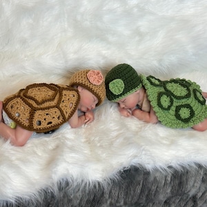 May include: Two babies dressed in handmade crochet turtle costumes, lying on a fluffy white surface. One costume is brown with a matching hat and a pink flower, the other is green with a green hat. The costumes resemble turtle shells.