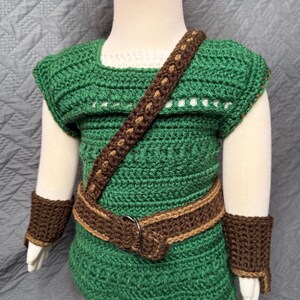 May include: A green and brown crochet outfit, including a tunic, belt, shoulder strap, and wrist cuffs. The tunic has a white neckline and is paired with a matching green hat. The outfit is designed for a child.