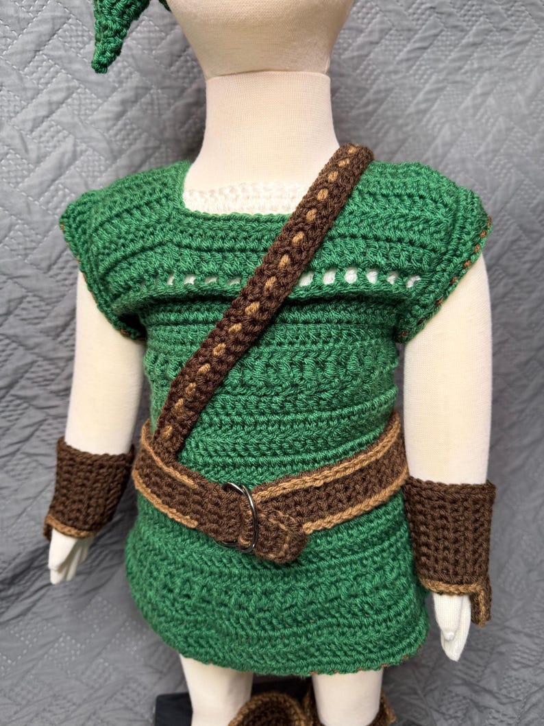 May include: A green and brown crochet outfit, including a tunic, belt, shoulder strap, and wrist cuffs. The tunic has a white neckline and is paired with a matching green hat. The outfit is designed for a child.