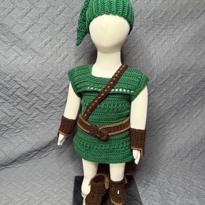 May include: A green crochet costume set, including a hat, tunic, belt, wrist cuffs, and boots. The tunic has a square neckline and a brown strap. The boots and cuffs are brown. The costume is displayed on a mannequin.