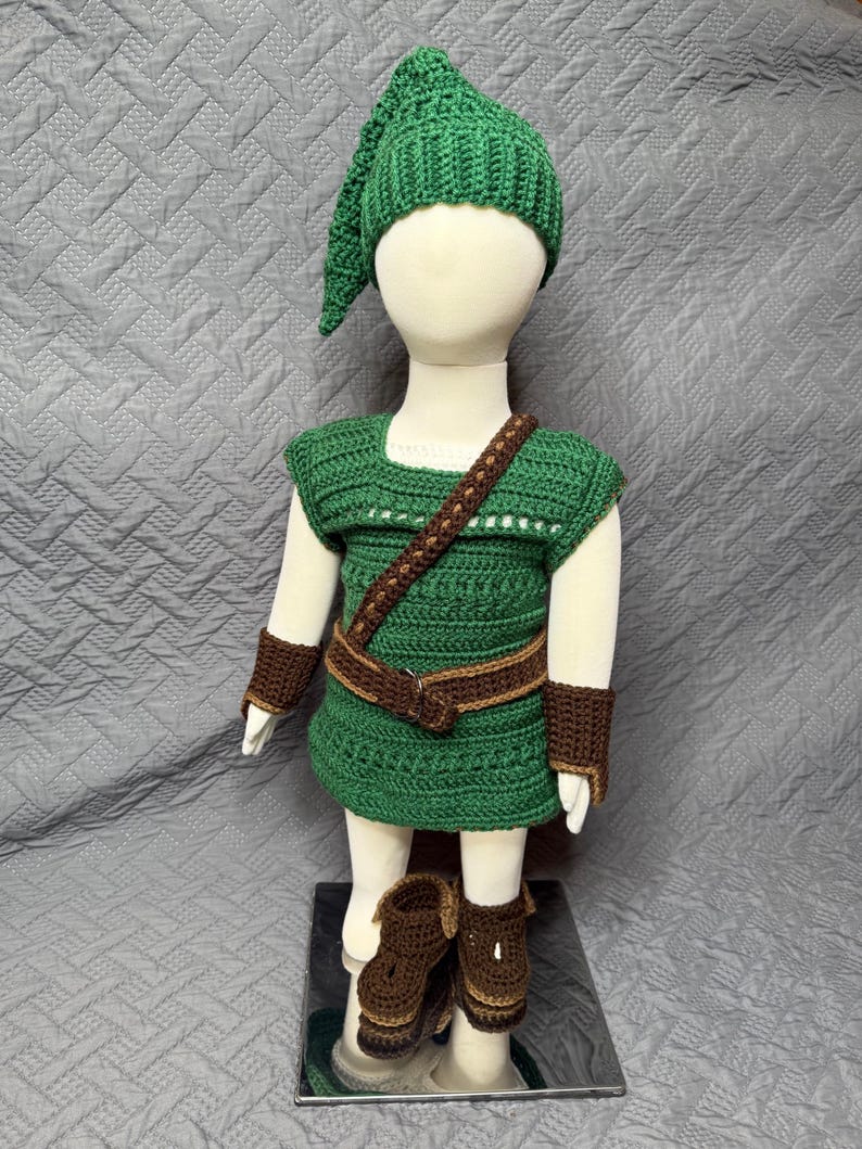 May include: A green crochet costume set, including a hat, tunic, belt, wrist cuffs, and boots. The tunic has a square neckline and a brown strap. The boots and cuffs are brown. The costume is displayed on a mannequin.