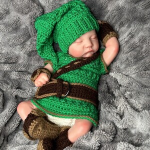 May include: A baby dressed in a handmade green and brown crochet outfit, including a hat, tunic, belt, arm cuffs, and booties. The baby is lying on a soft, gray blanket. The outfit resembles a character from a video game.