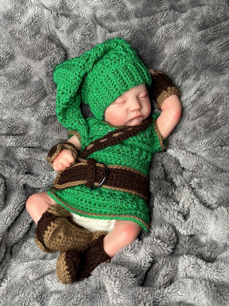 May include: A baby dressed in a handmade green and brown crochet outfit, including a hat, tunic, belt, arm cuffs, and booties. The baby is lying on a soft, gray blanket. The outfit resembles a character from a video game.