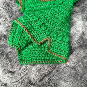 May include: A handmade, green crochet top with brown trim. The top has short sleeves and a square neckline. The crochet work is dense and detailed, showcasing the texture of the yarn. The garment is displayed on a soft, gray, textured surface.