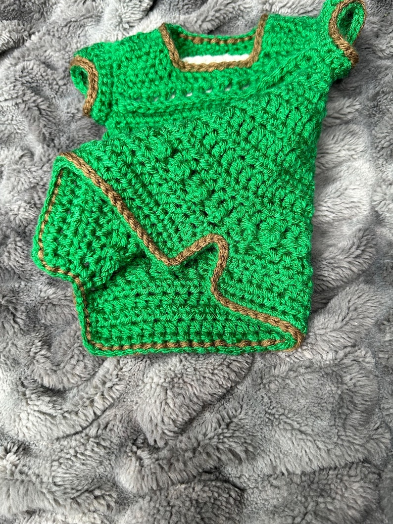 May include: A handmade, green crochet top with brown trim. The top has short sleeves and a square neckline. The crochet work is dense and detailed, showcasing the texture of the yarn. The garment is displayed on a soft, gray, textured surface.