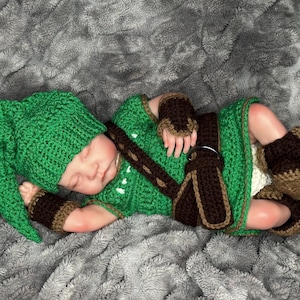 May include: A green crochet baby outfit, including a hat, tunic, wrist cuffs, and boots. The outfit has brown accents and a brown belt. The baby is lying on a gray, textured blanket.