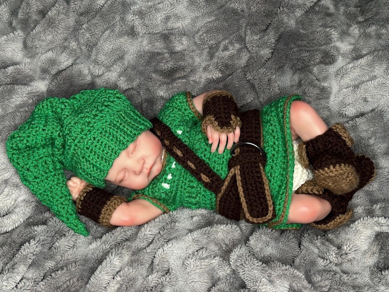 May include: A green crochet baby outfit, including a hat, tunic, wrist cuffs, and boots. The outfit has brown accents and a brown belt. The baby is lying on a gray, textured blanket.