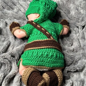 May include: A green and brown crochet baby outfit, including a hat, top, belt, mittens, and booties. The outfit is designed with a textured stitch and brown accents. The baby is lying on a gray, textured blanket.