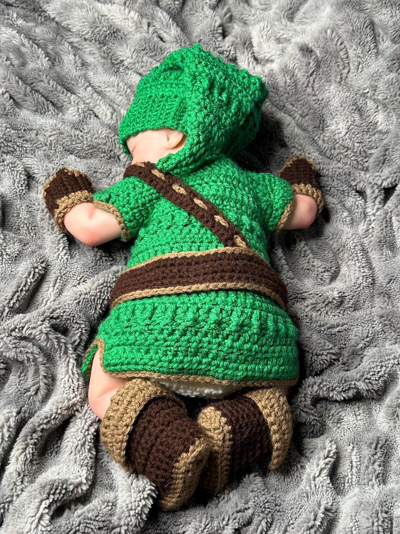 May include: A green and brown crochet baby outfit, including a hat, top, belt, mittens, and booties. The outfit is designed with a textured stitch and brown accents. The baby is lying on a gray, textured blanket.