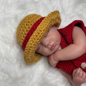 May include: A baby doll dressed in a handmade crochet outfit. The outfit features a yellow hat with a red band and a red vest. The doll is resting on a white, fluffy surface.