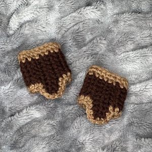 May include: Two handmade crochet cup sleeves. Each sleeve is brown with a tan trim. The sleeves are displayed on a soft, gray, textured surface.