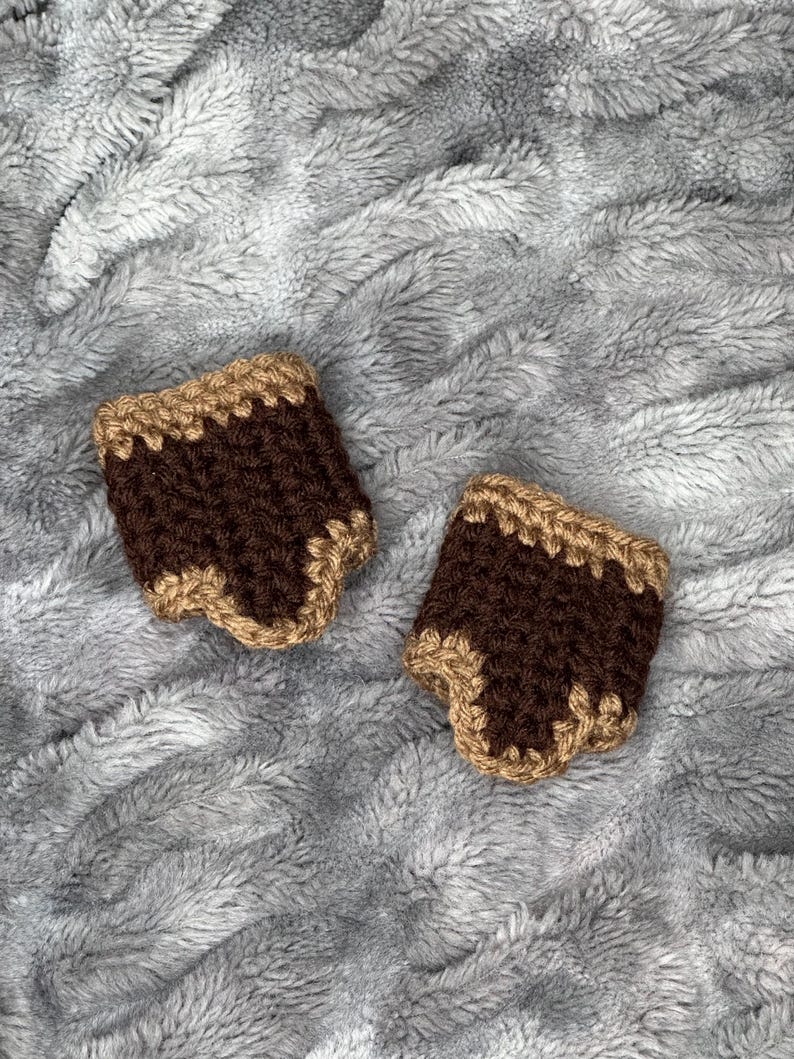 May include: Two handmade crochet cup sleeves. Each sleeve is brown with a tan trim. The sleeves are displayed on a soft, gray, textured surface.