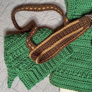 May include: A handmade crochet bag in shades of green, brown, and tan. The bag features a textured stitch pattern, a brown strap with tan accents, and a silver metal ring. The bag's design includes a leaf-like shape on the side.