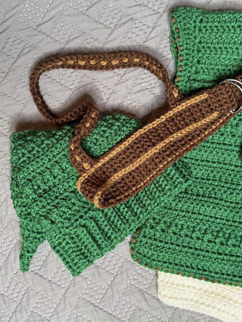 May include: A handmade crochet bag in shades of green, brown, and tan. The bag features a textured stitch pattern, a brown strap with tan accents, and a silver metal ring. The bag's design includes a leaf-like shape on the side.