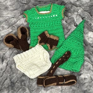 May include: A handmade crochet baby outfit in green, brown, and white. The set includes a top, hat, diaper cover, sandals, and suspenders. The green top and hat have brown trim. The sandals and suspenders are brown and tan.