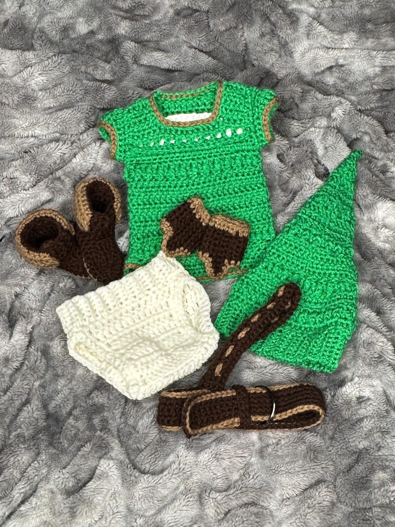 May include: A handmade crochet baby outfit in green, brown, and white. The set includes a top, hat, diaper cover, sandals, and suspenders. The green top and hat have brown trim. The sandals and suspenders are brown and tan.