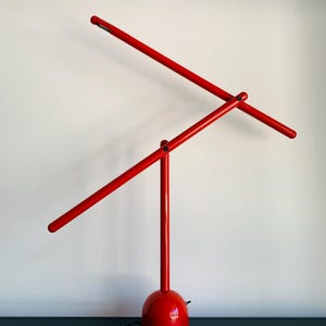 Puede incluir: Una lámpara de escritorio roja moderna con una base redondeada y dos brazos que se cruzan. La lámpara tiene un acabado brillante y un diseño minimalista, con un cable de alimentación negro que sale de la base.