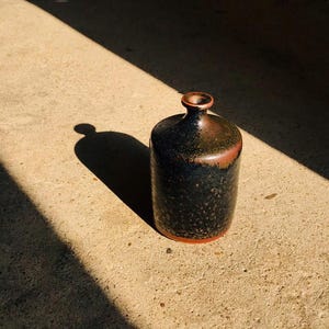 Puede incluir: Un jarrón cilíndrico de cerámica con cuello estrecho y esmalte moteado negro y marrón. El jarrón tiene un borde y una base de color marrón rojizo, sobre un fondo neutro.