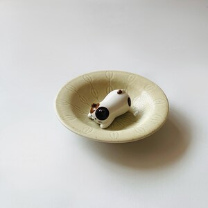 以下が含まれることがあります： 茶色と黒の模様が入った小さなセラミック製の猫の置物が、ベージュの模様の入ったお皿の中にあります。猫は白で、茶色の耳と黒い斑点があります。お皿には葉のような模様があります。