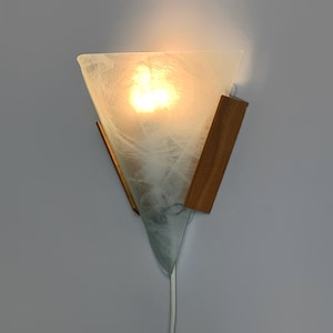 May include: A triangular wall sconce featuring a translucent white glass shade with a marbled texture, illuminated from within by a warm light. The shade is secured by two vertical wooden side panels and includes a white power cord extending downward.