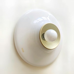 Op de afbeelding: Een witte, moderne wandlamp met een afgeronde, schotelvormige kap. De lamp heeft een goudkleurige binnenring en een witte lamp. De lamp is op een witte muur gemonteerd.