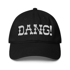 May include: Black baseball cap with the word "DANG!" embroidered in white across the front. The cap has a curved brim and a fabric-covered button on top. The text is in a bold, stylized font.