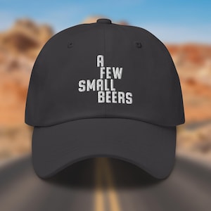 May include: A dark gray baseball cap with the embroidered text "A FEW SMALL BEERS" in white. The cap has a curved brim and a button on top. The background is a blurred image of a road and desert landscape.