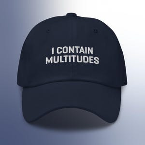 Op de afbeelding: Marineblauwe baseballcap met de geborduurde tekst "I CONTAIN MULTITUDES" in wit. De cap heeft een gebogen klep en een knoop aan de bovenkant. Een klassiek, casual accessoire.