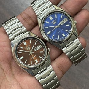 Op de afbeelding: Twee Seiko automatische horloges met roestvrijstalen banden. Eén horloge heeft een bruine wijzerplaat, de andere een blauwe. Beide horloges tonen de datum en dag. De horloges worden op een hand getoond.