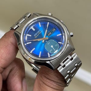 May include: A stainless steel Seiko Speedtimer watch with a vibrant blue dial. The watch features a day and date display, along with sub-dials and a silver-tone bracelet. The watch is held in a hand.