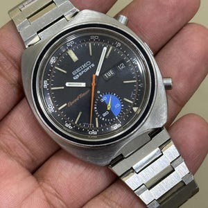 May include: A vintage Seiko Sports SpeedTimer watch with a black dial, silver case, and a stainless steel band. The watch face displays the day and date, with orange and blue accents. The watch has a chronograph function.