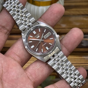 Może przedstawiać: Automatyczny zegarek na rękę Seiko ze stali nierdzewnej z brązową tarczą. Zegarek posiada wyświetlacz dnia i daty oraz srebrny metalowy pasek. Zegarek jest trzymany w dłoni.