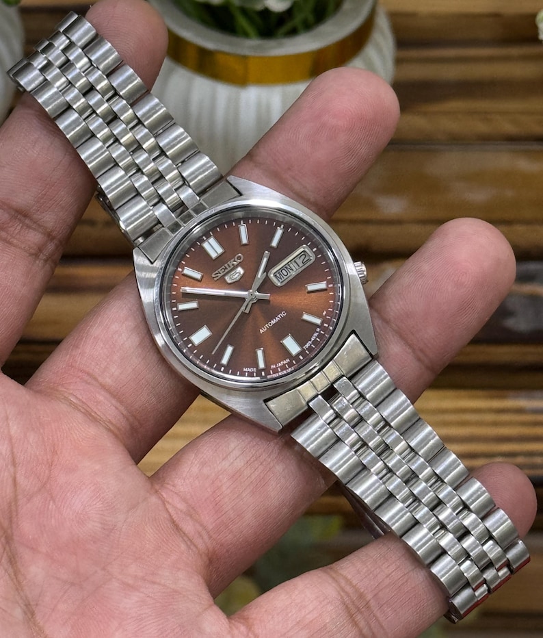 Może przedstawiać: Automatyczny zegarek na rękę Seiko ze stali nierdzewnej z brązową tarczą. Zegarek posiada wyświetlacz dnia i daty oraz srebrny metalowy pasek. Zegarek jest trzymany w dłoni.