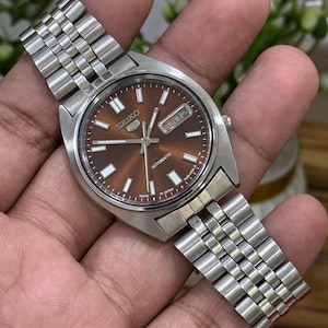 Może przedstawiać: Automatyczny zegarek Seiko 5 ze stali nierdzewnej z brązową tarczą. Zegarek posiada wyświetlacz dnia i daty oraz srebrną metalową bransoletę. Zegarek jest trzymany w dłoni.